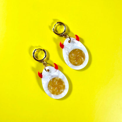 A pair of novelty earrings shaped like fried eggs, featuring white eggshells with gold-colored rims, yellow yolks, and small red accents for the beaks and feet, shown on a bright yellow background.