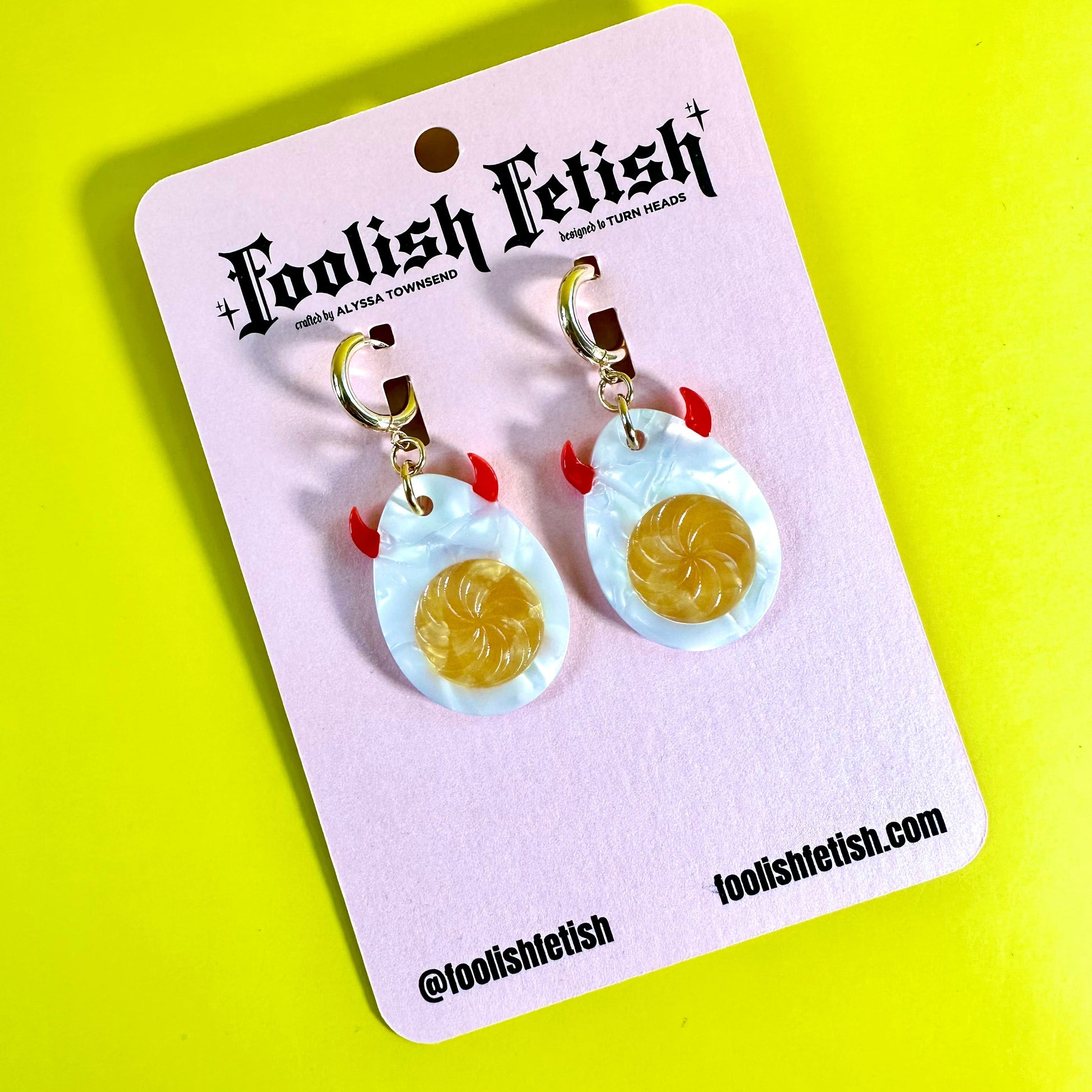 A pair of dangling earrings displayed on a pink card. Each earring has a gold fishhook clasp, a small red loop, and a light blue marbled oval charm with a yellow swirl in the center.