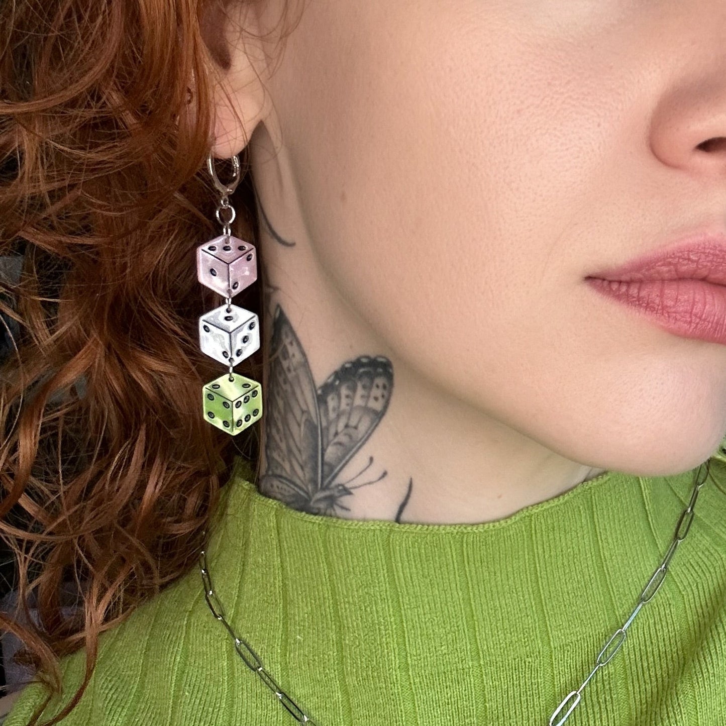 A close-up lifestyle shot of a person with curly red hair wearing a green ribbed top, a silver necklace with a black and green winged pendant, and dangling geometric earrings.