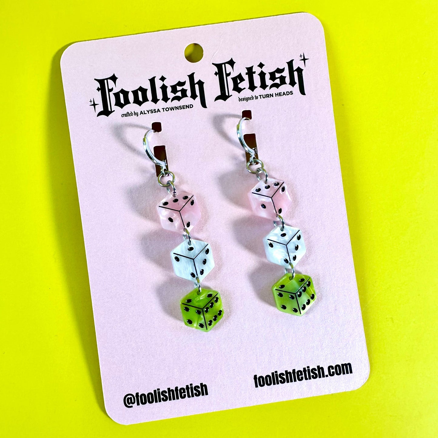 A pair of dangling dice earrings displayed on a white branded card against a bright yellow background. The earrings feature translucent pink, white, and lime green dice with black pips, and the card shows the brand name 'Foolish Fetish'.