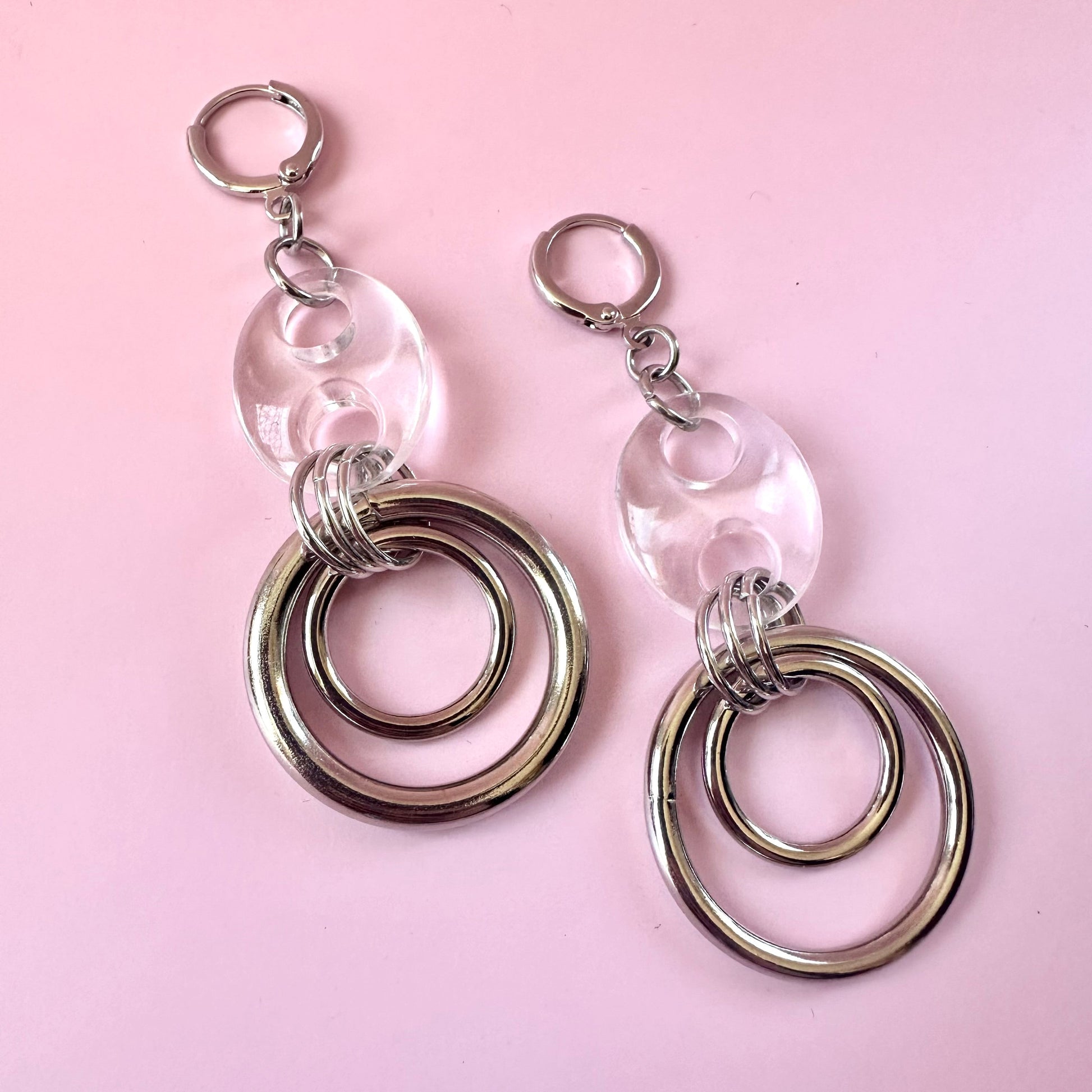 A pair of silver-toned dangle earrings with clear round acrylic discs and stacked concentric metal rings, displayed on a solid pink background.