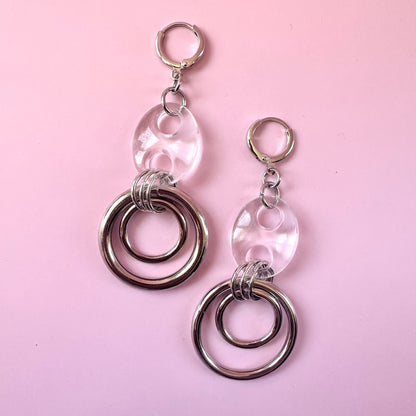 A pair of handmade dangle earrings featuring clear, oval-cut glass beads with silver-toned metal hoops and jump rings, displayed on a solid pink background.