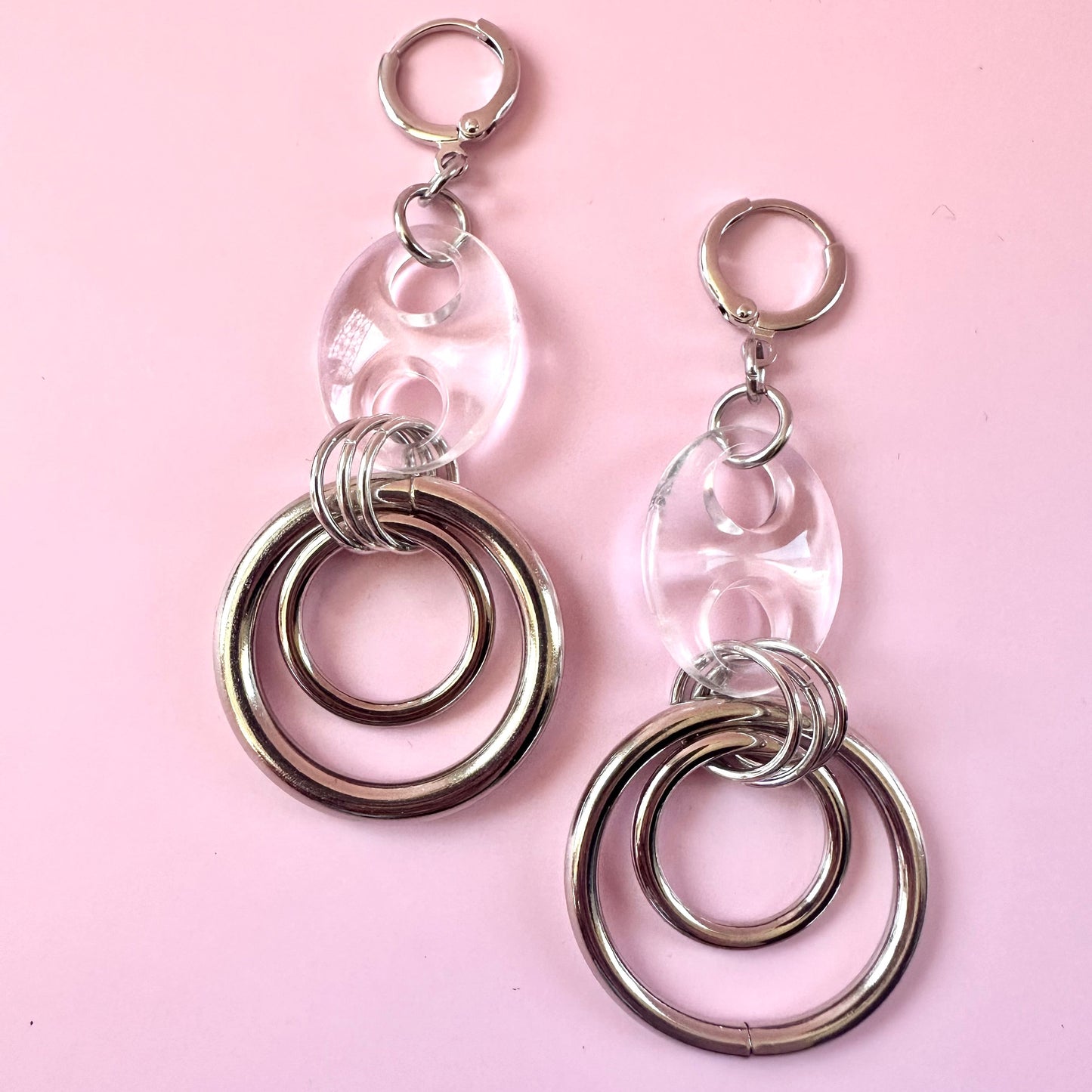 A pair of silver-toned dangle earrings with clear oval resin pendants and double-ring metal charms, displayed on a solid light pink background.