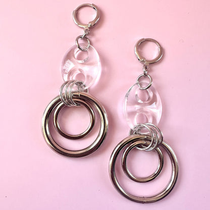 A pair of silver-toned dangle earrings with clear oval resin pendants and double-ring metal charms, displayed on a solid light pink background.