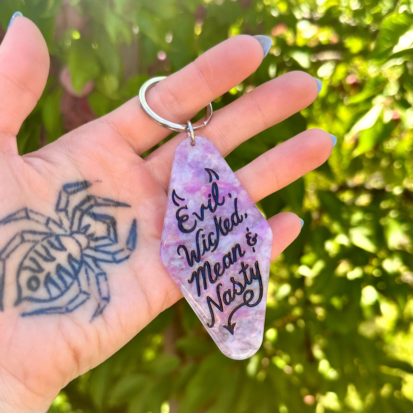 A hand holding a purple and lavender stone keychain engraved with 'Evil. Wicked, Mean, and Nasty