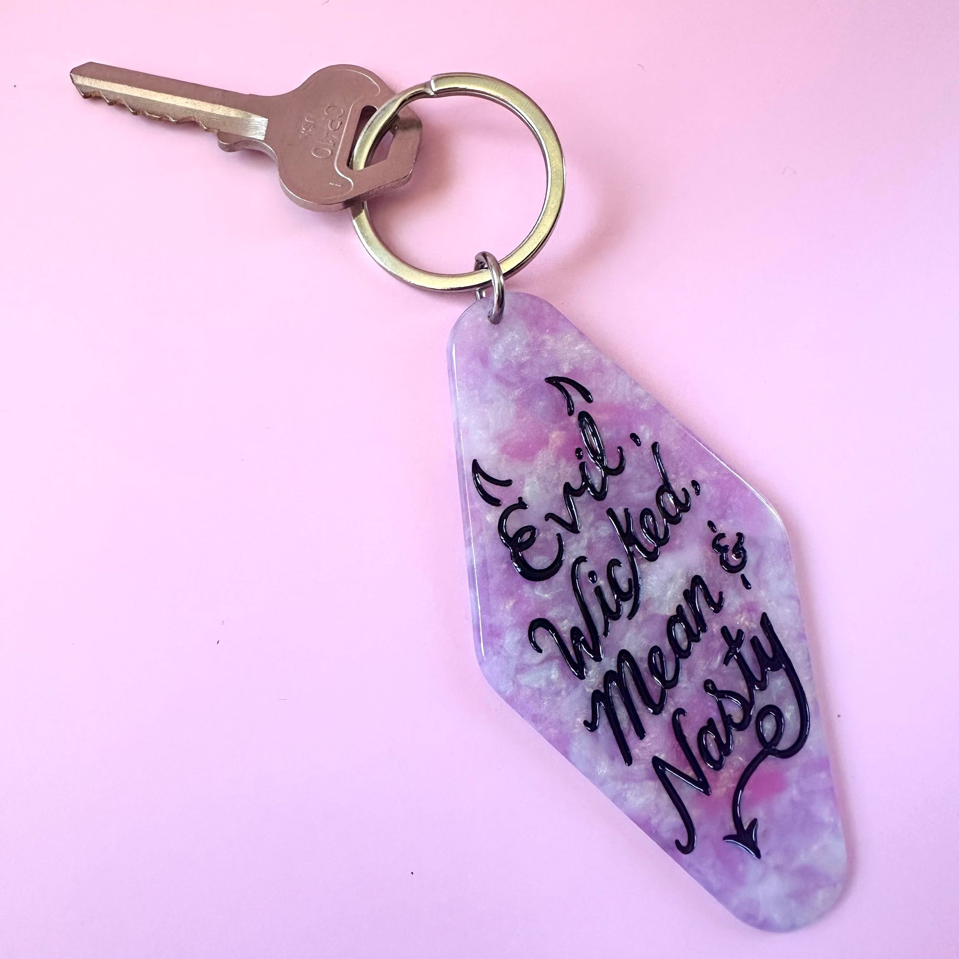 A keychain with a purple and pink marbled charm featuring the text 'Evil, Wicked, Mean & Naughty' in black script, attached to a silver metal keyring with a key on a solid light pink background.