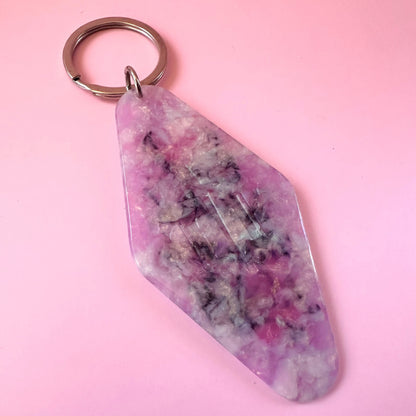 A pink and purple marbled crystal keychain with a silver metal split ring, shown on a solid pink background.