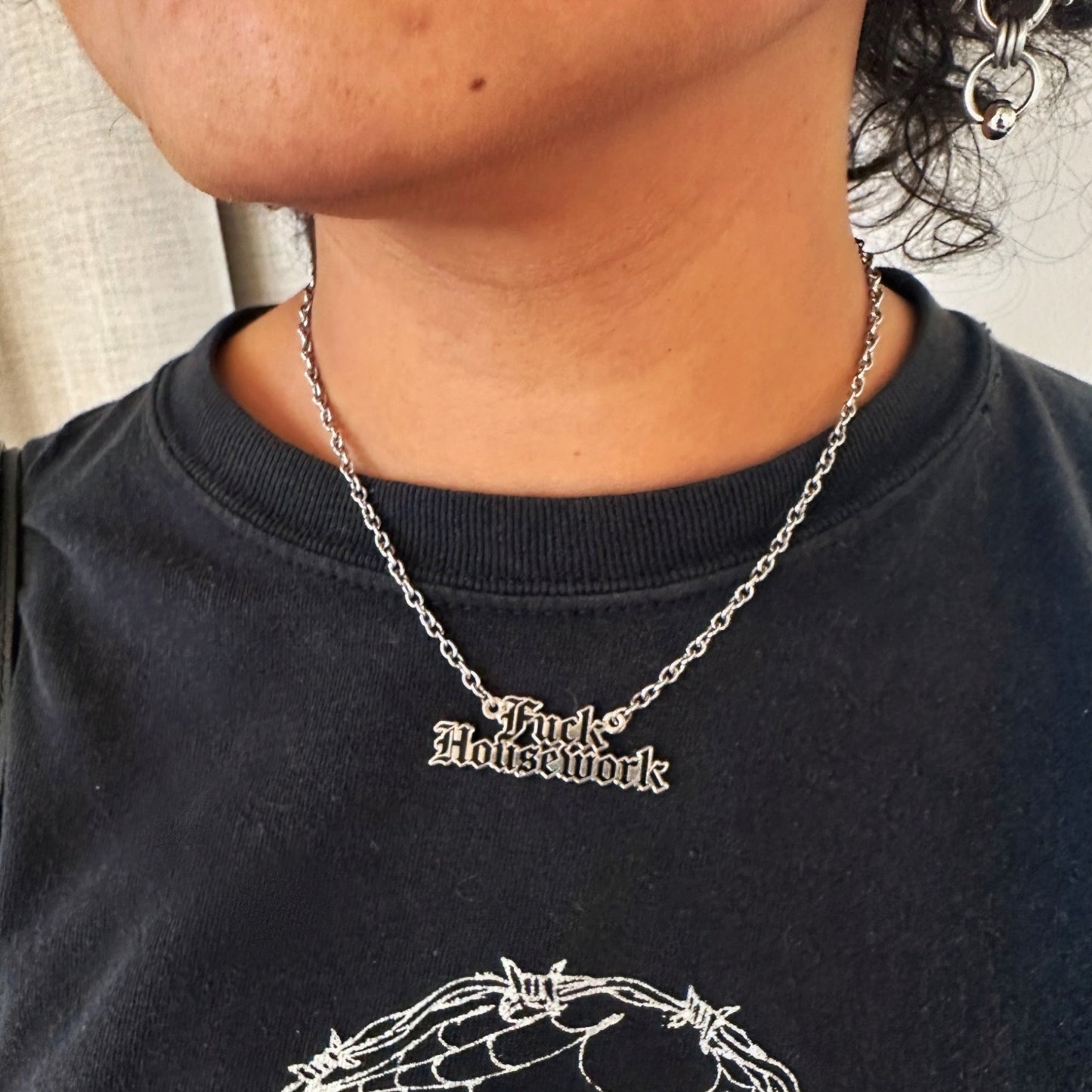 Person wearing a black shirt with a spider web design and a necklace with 'Fuck Housework' pendant.