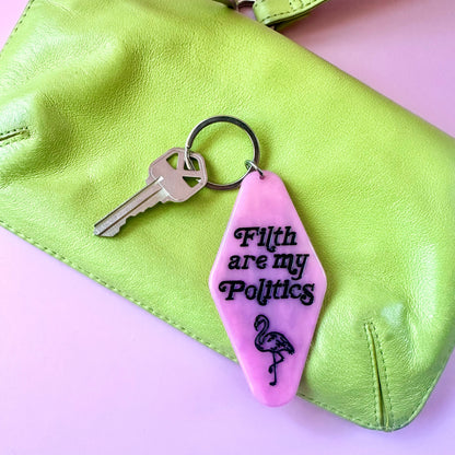 A pink acrylic keychain with the text 'Filth are my Politics' and a flamingo graphic, attached to a key ring and displayed on a lime green leather clutch against a light pink background.