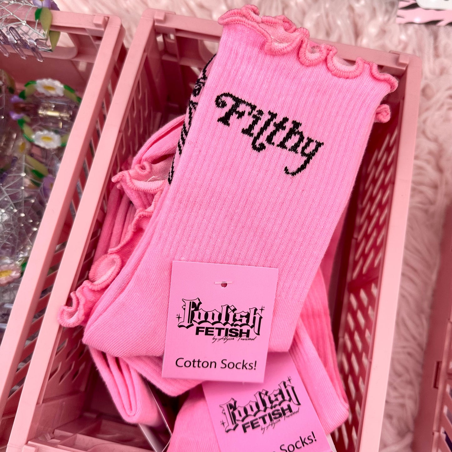 Pink cotton socks with black gothic-style text reading 'Fitsly' and 'Foot Fetish' on tags, displayed in a pink basket on a white faux-fur surface.