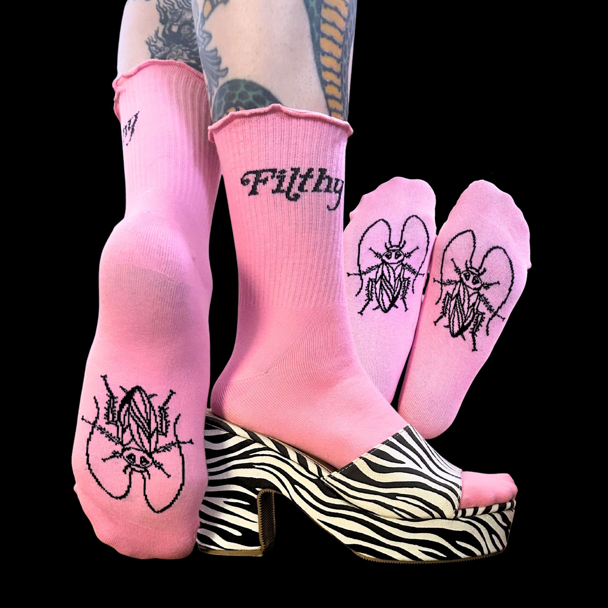 Pink crew socks with black embroidered gothic-style text and insect illustrations, shown on a model's feet paired with zebra-print platform heels against a black background.