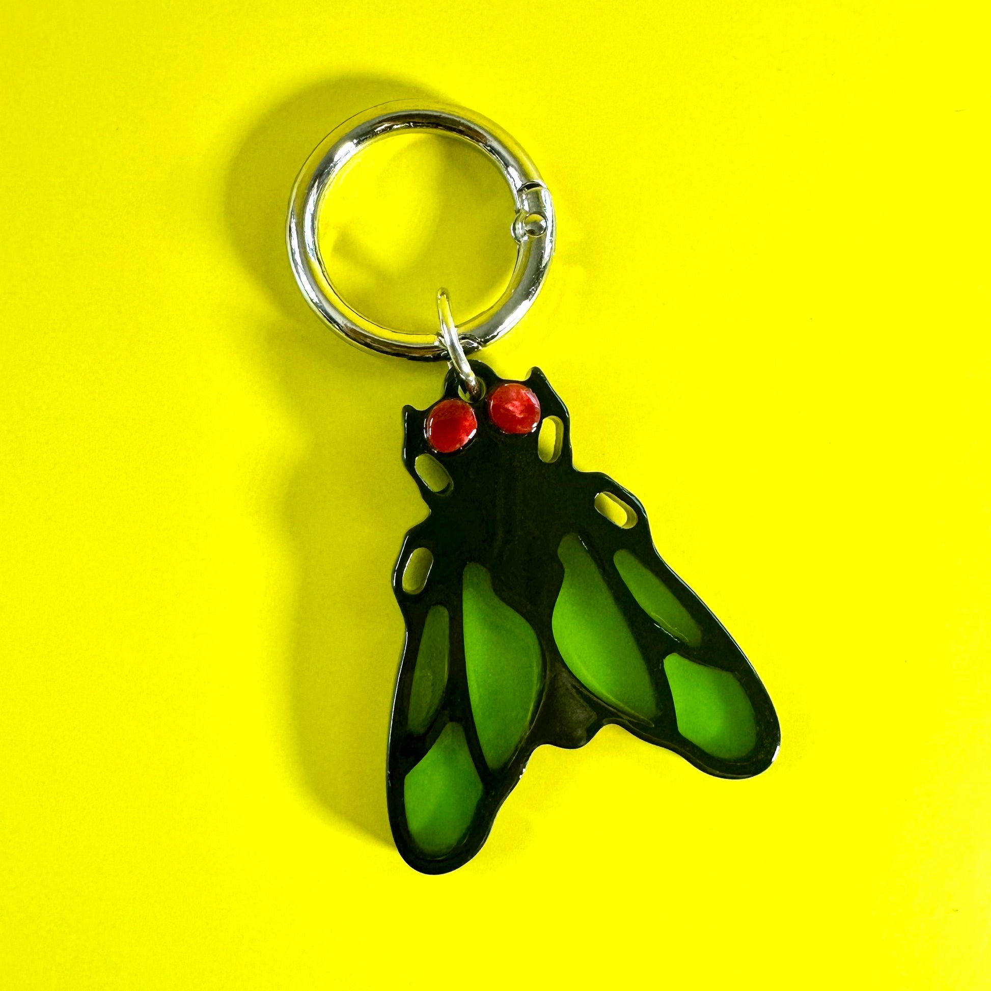 A green and black winged keychain charm with small red eyes, attached to a silver split ring, set against a solid bright yellow background.