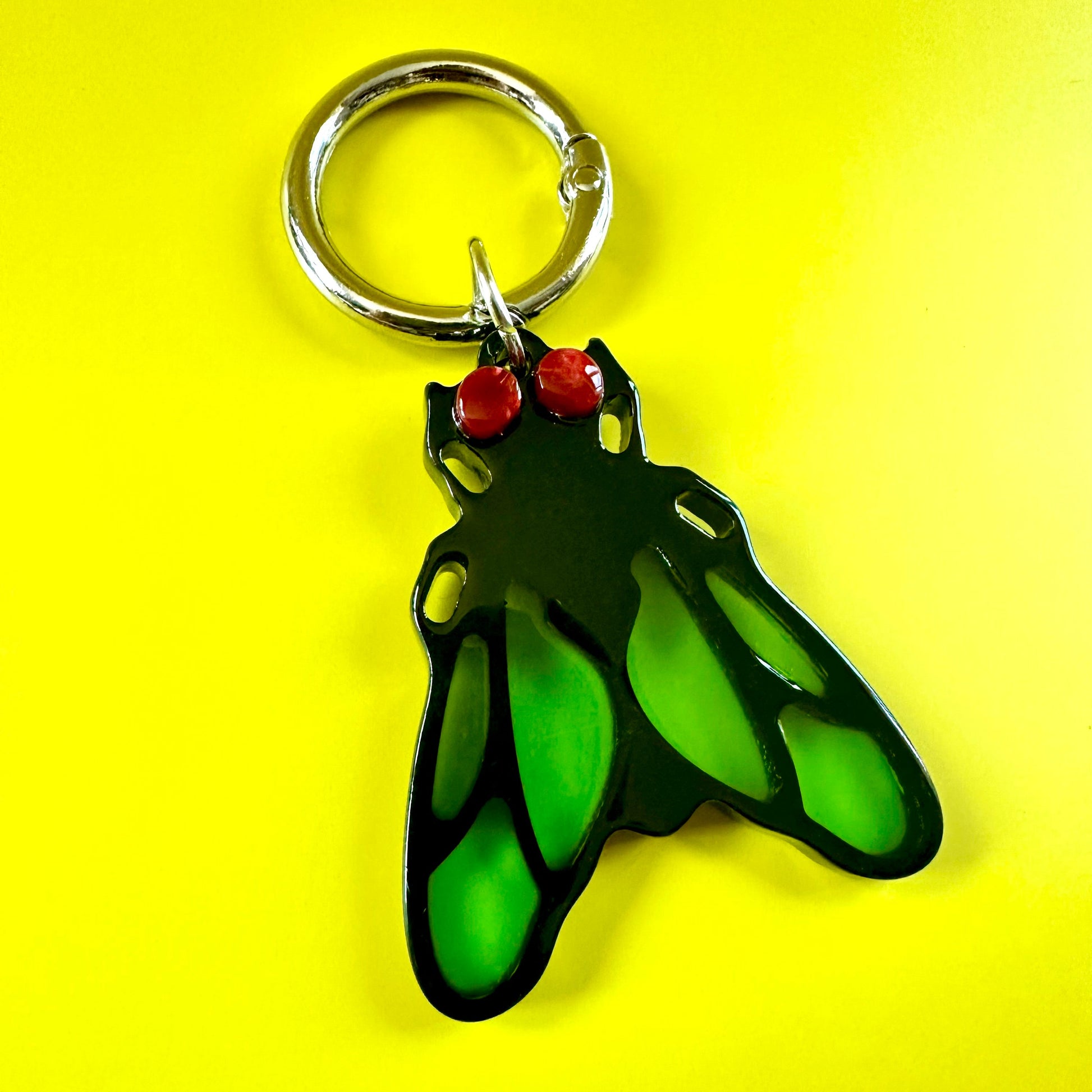 A green and black winged insect keychain charm with red eyes, attached to a silver metal split ring, set against a solid yellow background.