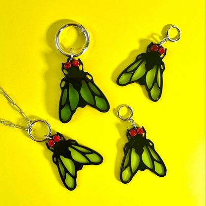 Three insect-themed jewelry charms or pendants with silver key rings and chain, featuring green wings with black outlines and red heads, arranged on a bright yellow background.