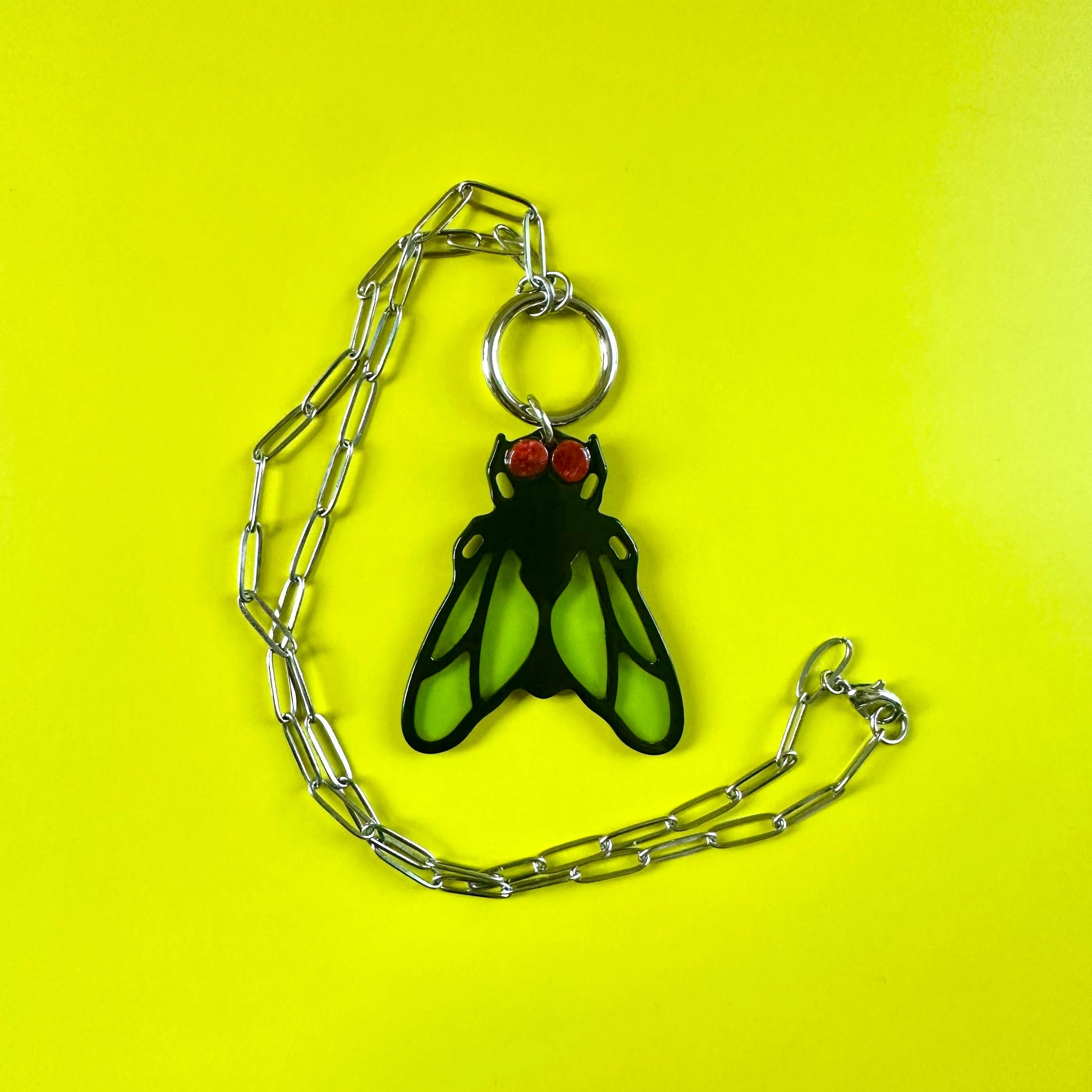 A gothic insect pendant necklace featuring a black and neon green fly design with red eyes, suspended on a silver paperclip chain against a bright yellow background.