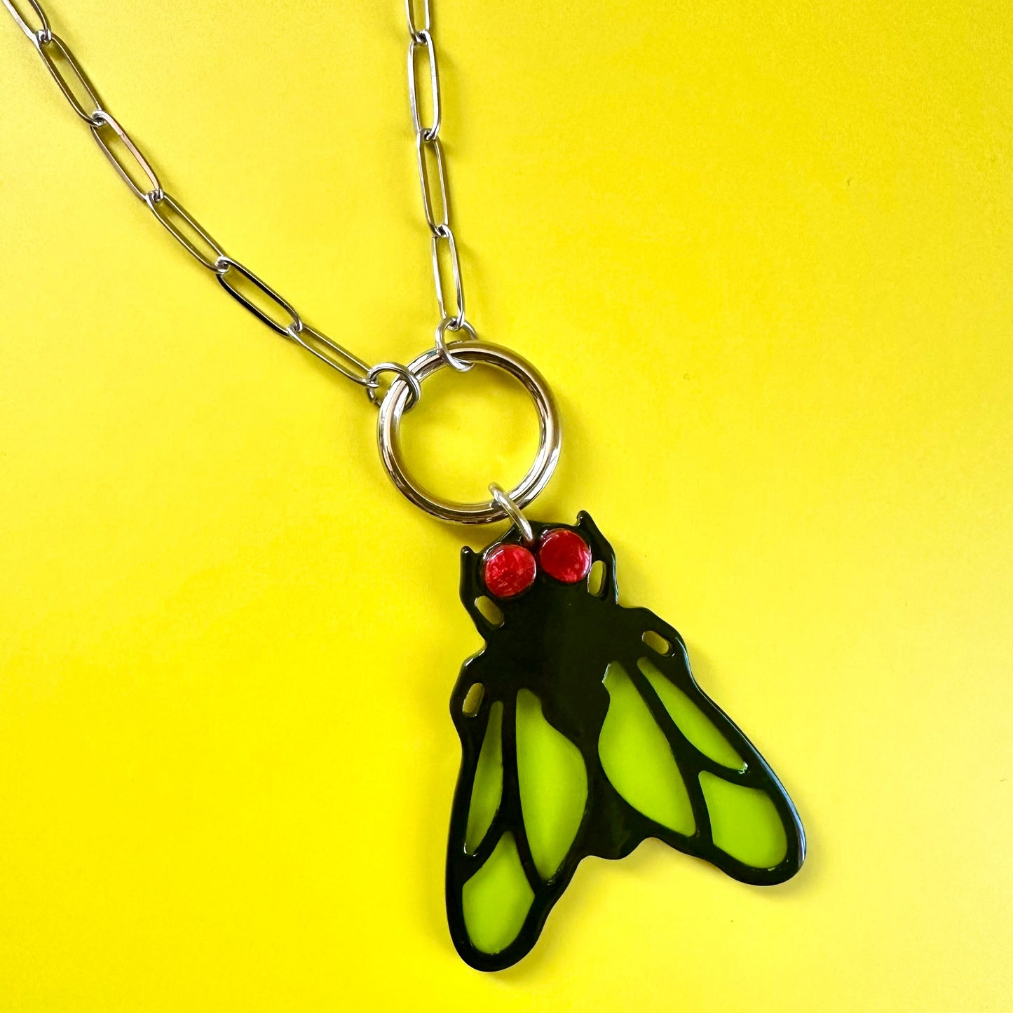 A silver paperclip chain necklace with a circular ring and a cutout pendant shaped like a fly, featuring bright neon green wings and two small red eyes, displayed on a solid yellow background.