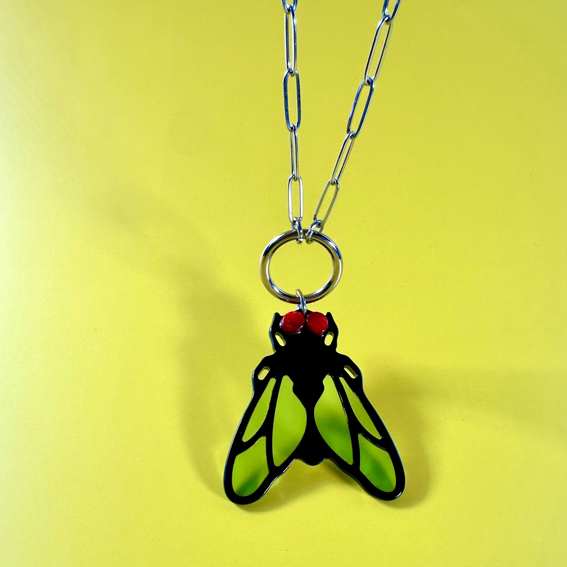 A pendant necklace featuring a stylized insect or fly design with bright lime green wings outlined in black, hanging from a silver link chain against a solid pale yellow background.
