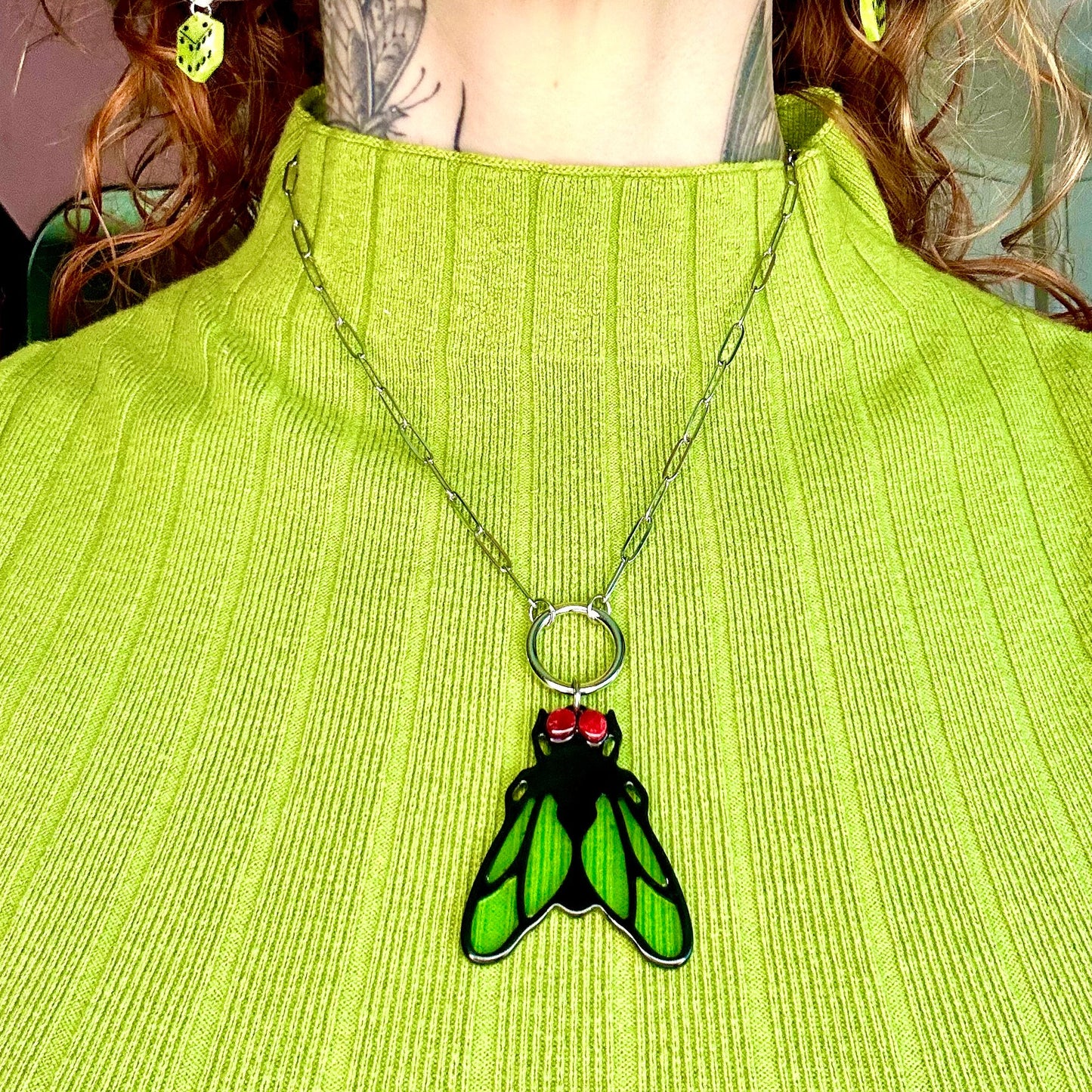A close-up of a person wearing a green and black enamel fly pendant necklace over a lime green ribbed turtleneck sweater.