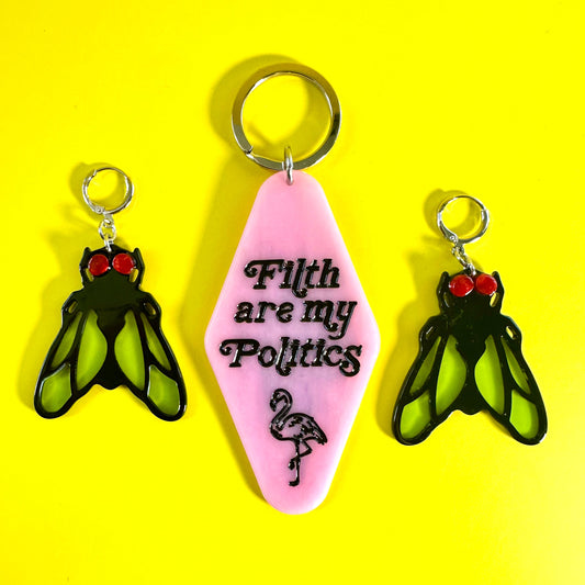 A pink guitar-shaped keychain with the text 'Filth are my Politics' and a flamingo illustration, shown with two matching winged keychain charms on a bright yellow background.