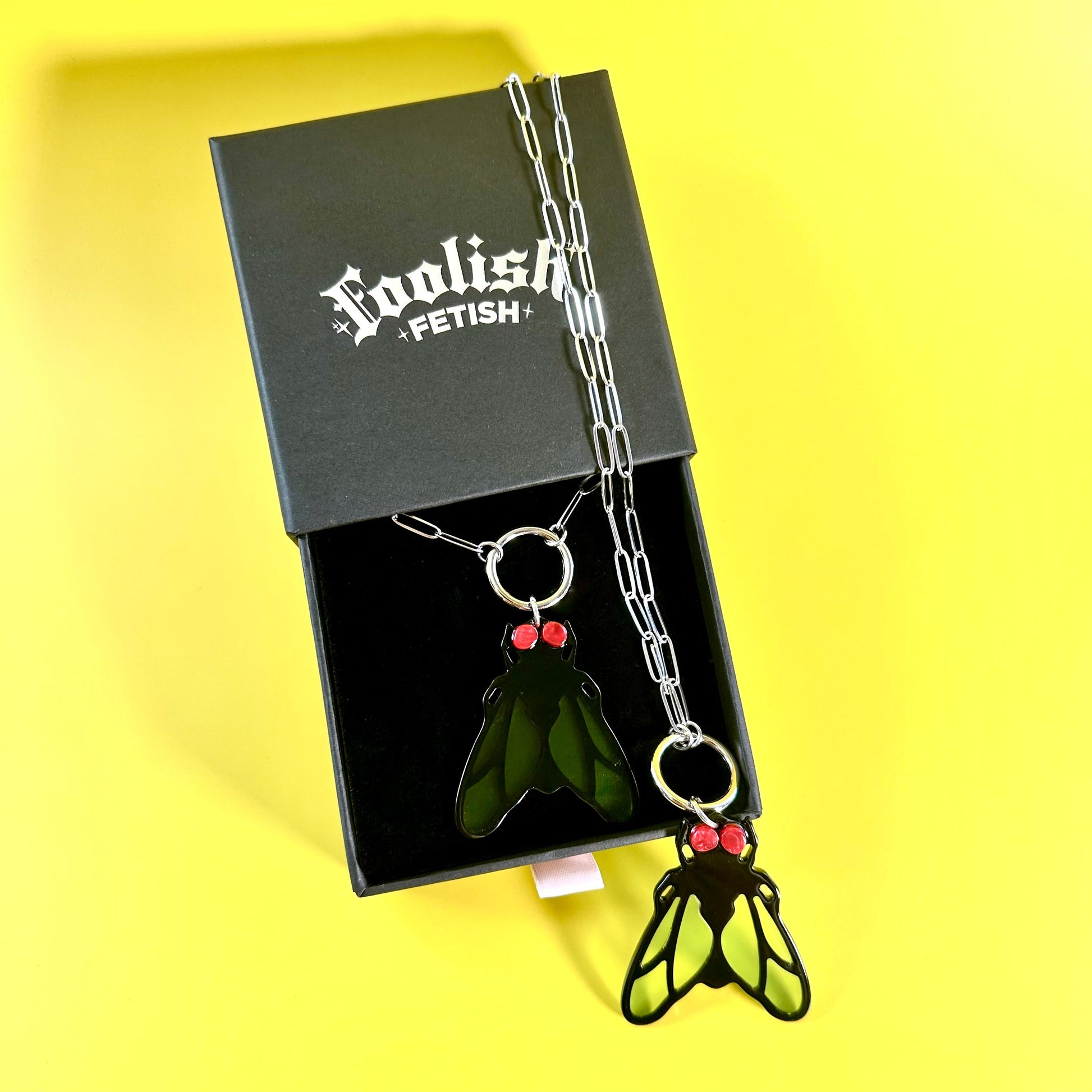 A novelty pendant necklace displayed in an open black box on a bright yellow background. The necklace has a silver chain and two insect-themed pendants: one shaped like a green leaf with red berry accents, and one shaped like a black-and-green winged insect with red berry accents.