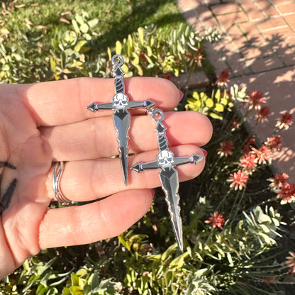 Silver dagger-shaped sword earrings  with skull detail held in a hand with a garden background