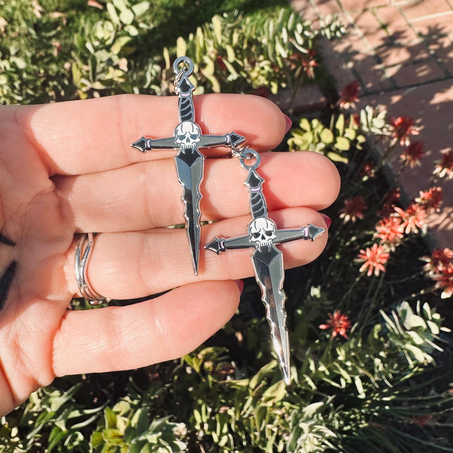 Hand holding two silver sword-shaped pendants with skull designs against a natural background.