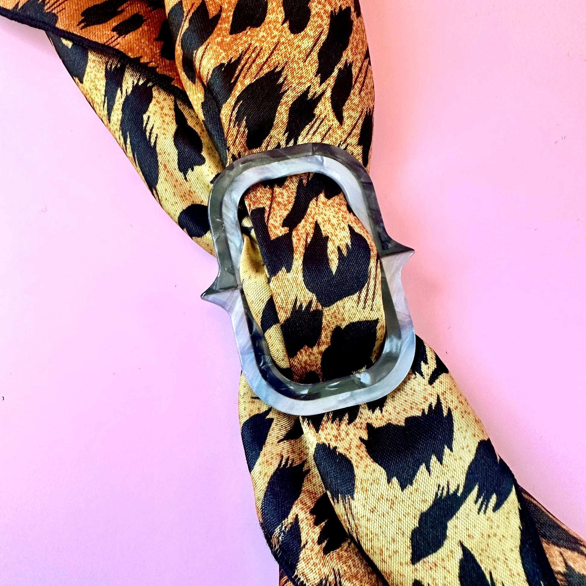 A leopard print scarf tied in a knot with a silver metal ring, set against a solid pink background.