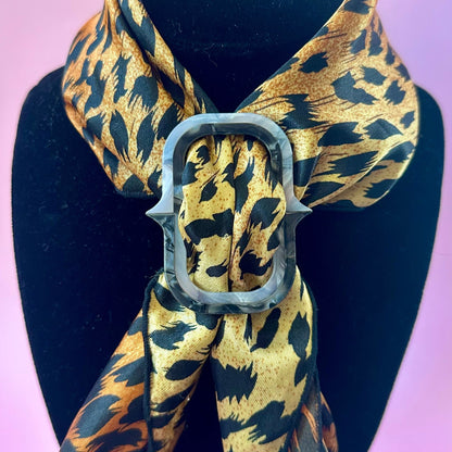 A leopard print scarf with a large silver buckle, displayed on a black mannequin bust against a soft pink background.