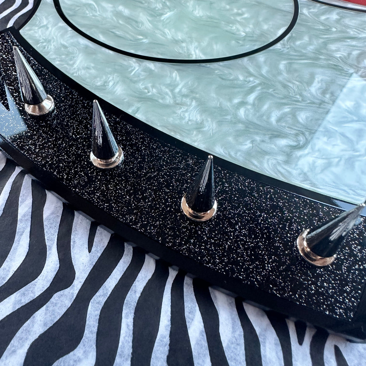 A close-up of a decorative tabletop or tray featuring a stylized cartoon cat face in pale mint green, red lips, and a black glitter border with metallic spike accents, resting on a black-and-white zebra print surface.
