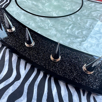 A close-up of a decorative tabletop or tray featuring a stylized cartoon cat face in pale mint green, red lips, and a black glitter border with metallic spike accents, resting on a black-and-white zebra print surface.