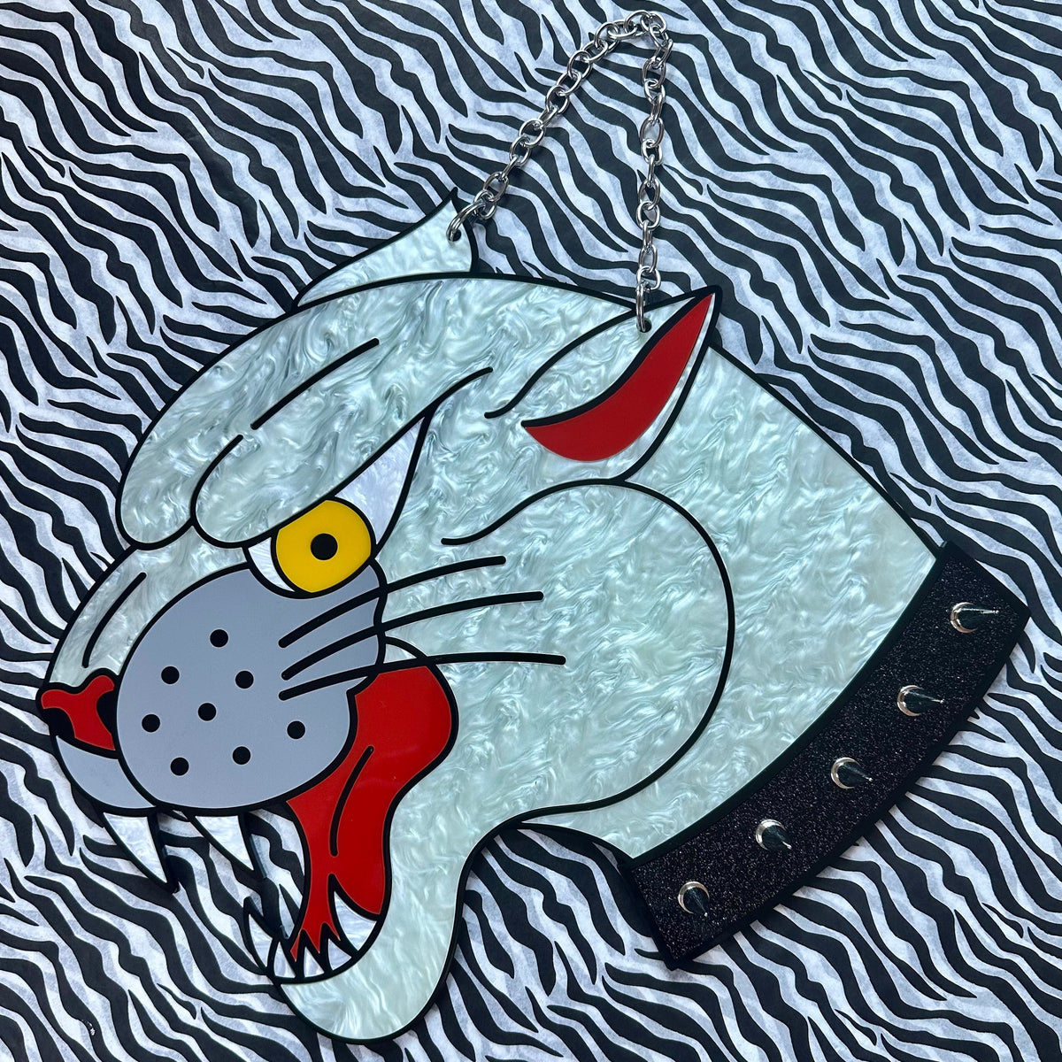 A handbag featuring a stylized panther head patch in light blue, red, yellow, and gray, with a black studded collar detail and a silver chain strap, set against a black and white zebra-print fabric background.