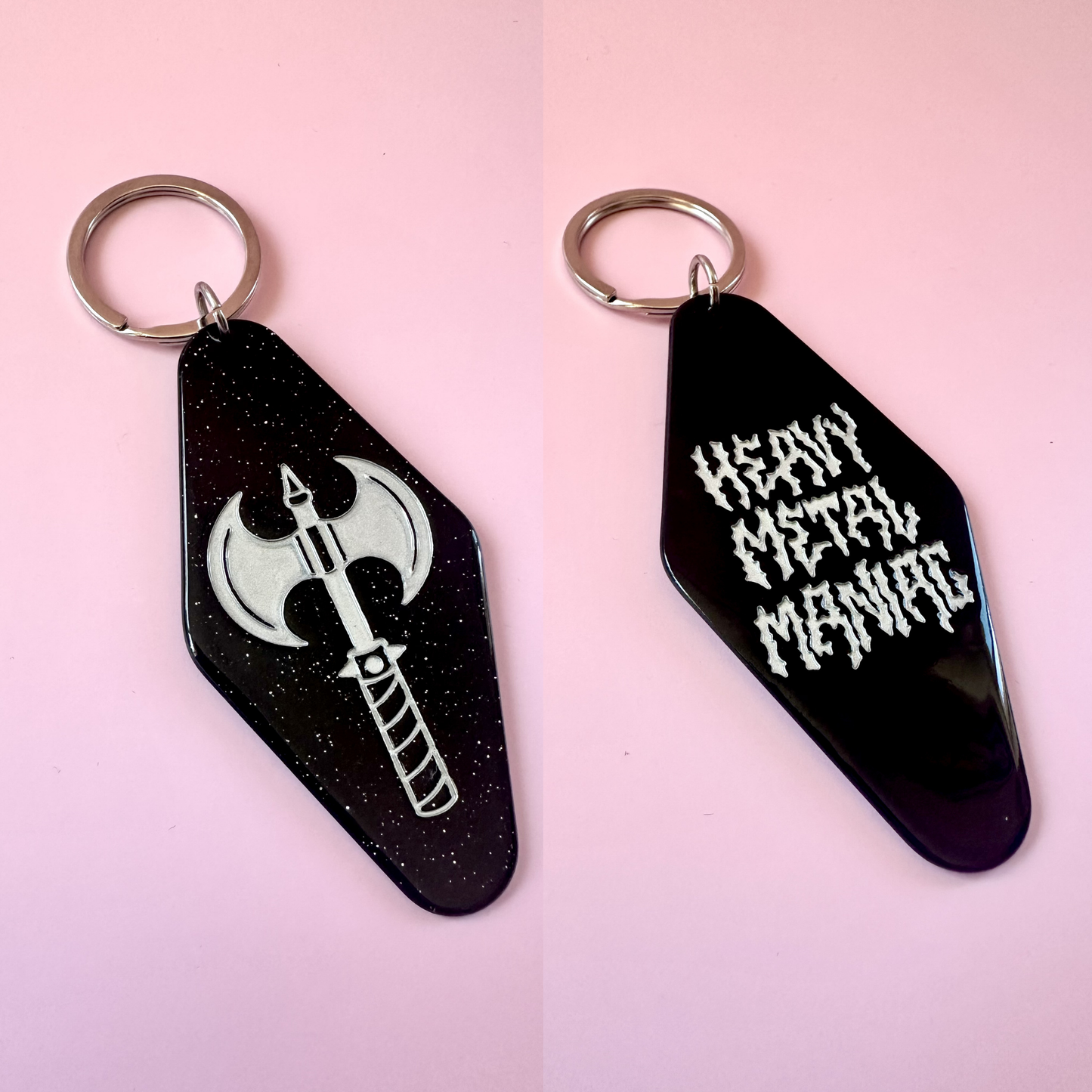 Two black gothic-shaped keychains with silver key rings on a pink background. One keychain features a white axe graphic, and the other features the text 'HEAVY METAL MANIC' in a white gothic font.