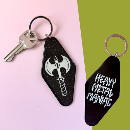 Two black hotel keychains with metal key rings are displayed on a split pink and green background. One keychain features a silver axe graphic, and the other features the text 'HEAVY METAL MANIC' in a white gothic font.