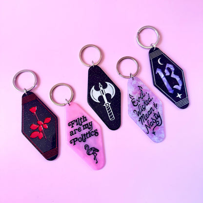 Four hotel style keychains with silver keyrings, arranged on a light pink background. Each keychain has a different gothic-themed design: one black with a red rose and heart, one pink with the text 'F*ck are my Politics' and a snake, one black with a white axe, and one lavender-purple with the number 13 and a crescent moon.
