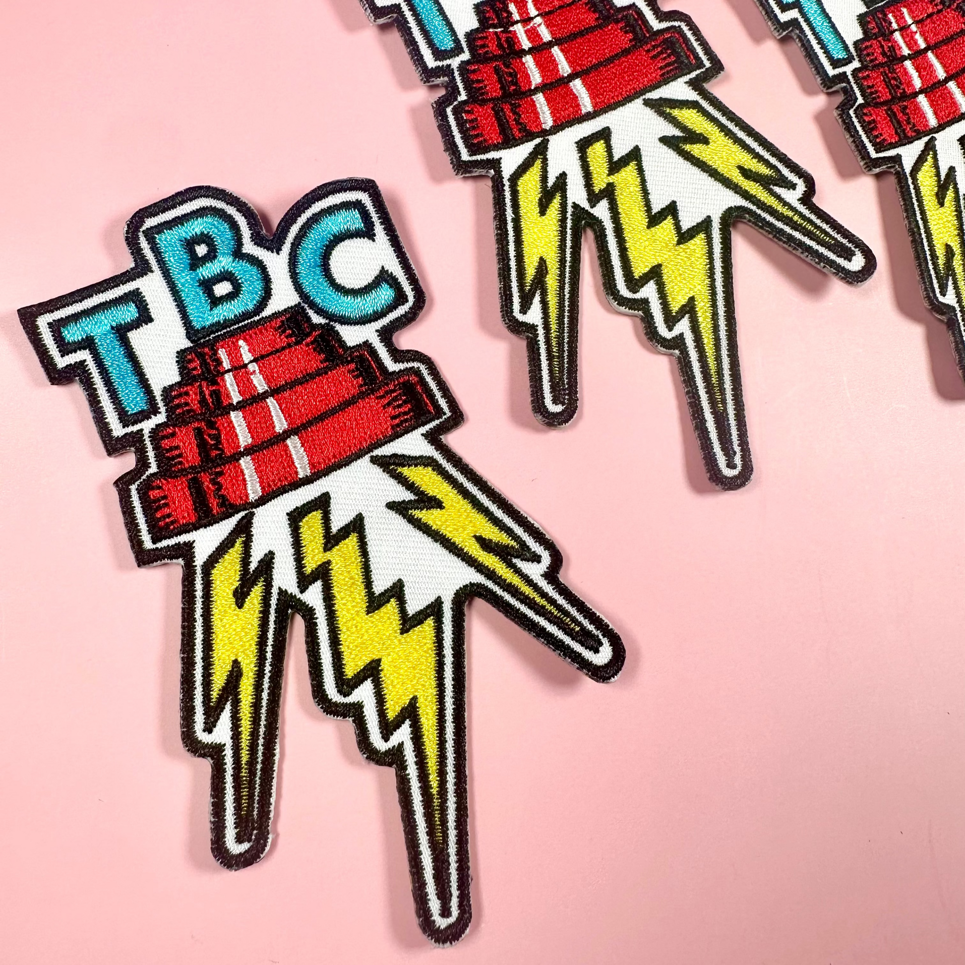 Three 'TBC' logo patches with lightning bolts on a pink background