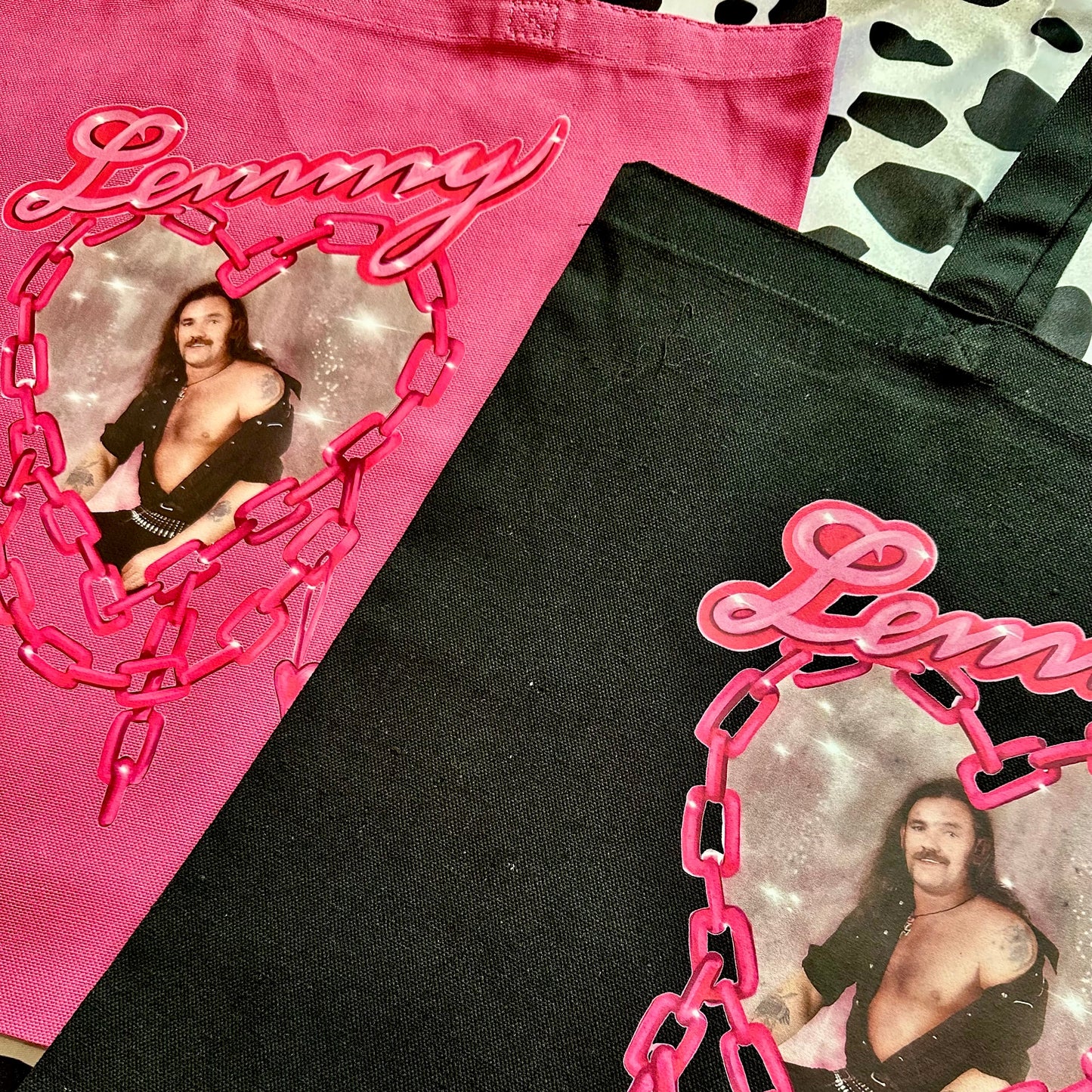 Two tote bags shown in a flat-lay arrangement: one bright pink bag and one black bag, both featuring a large pink graphic with the word "Lemmy" in script, a heart-shaped chain design, and a printed photo of Lemmy Knight.