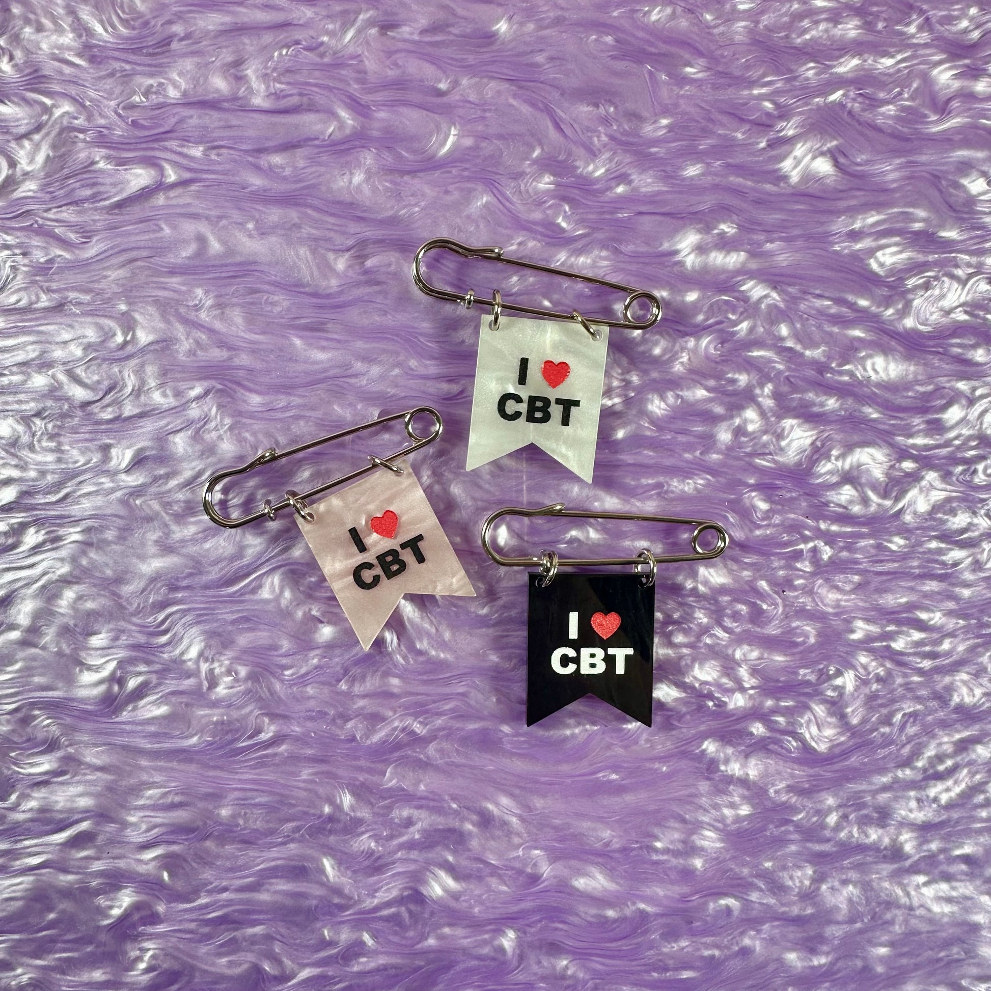 Three safety pin badges with 'I ❤️ CBT' text on a purple textured background