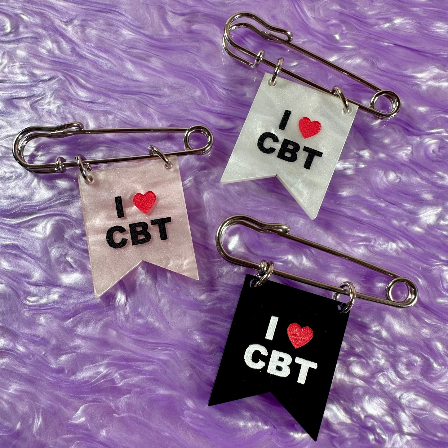 Three safety pin brooches with 'I ❤️ CBT' text on a purple textured background