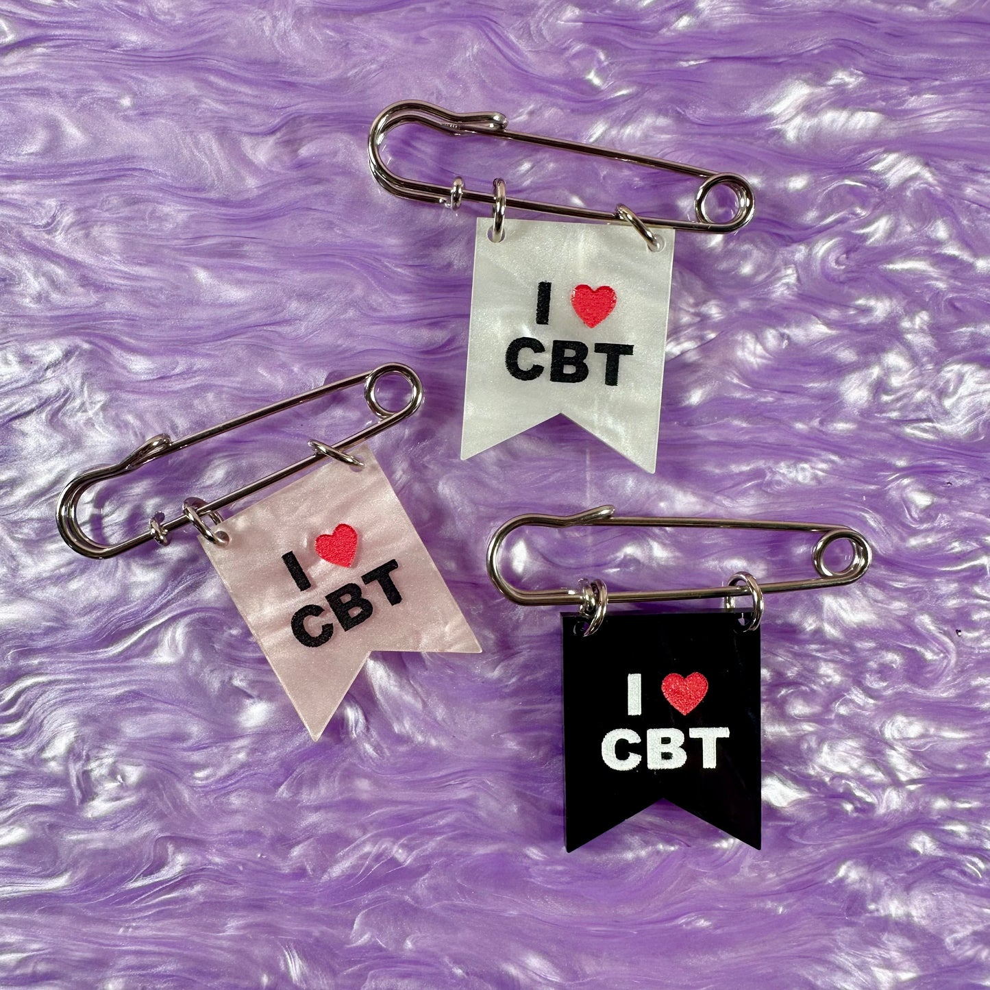 Three safety pin brooches with 'I ❤️ CBT' text on a purple textured background