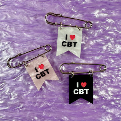 Three safety pin brooches with 'I ❤️ CBT' text on a purple textured background