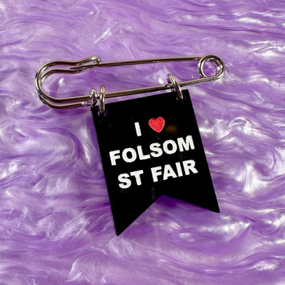 Black flag-shaped keychain with 'I ❤️ FOLSOM ST FAIR' text on a purple textured background