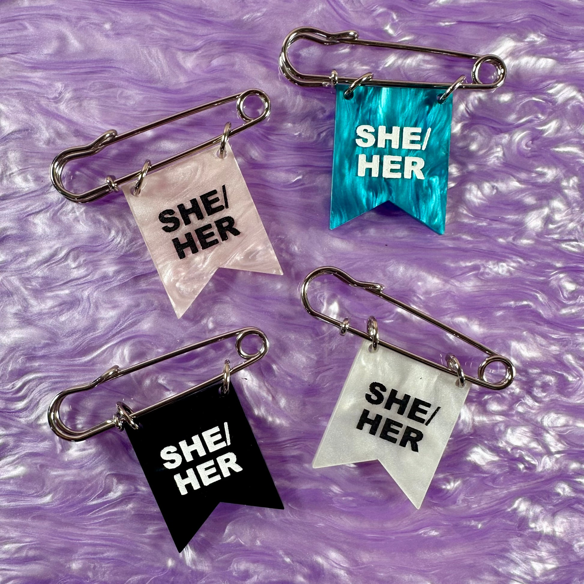Four 'SHE/HER' flags on safety pins against a purple textured background