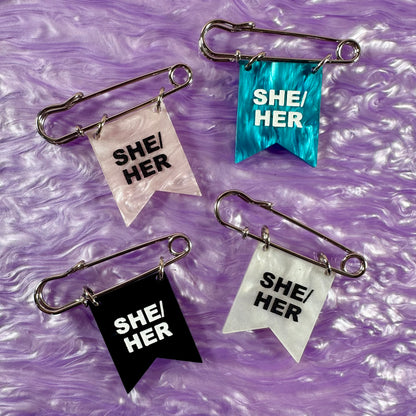 Four 'SHE/HER' flags on safety pins against a purple textured background