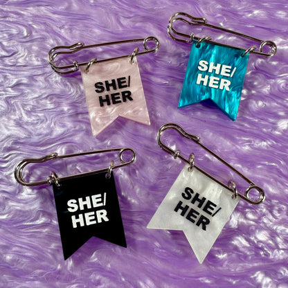 Four 'SHE/HER' pride pins on a purple textured background