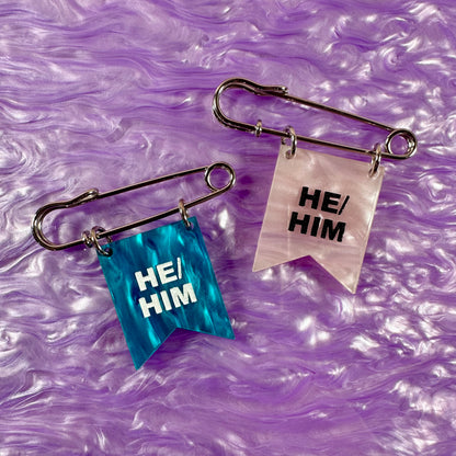 Two safety pins with 'HE/HIM' labels on a purple textured background
