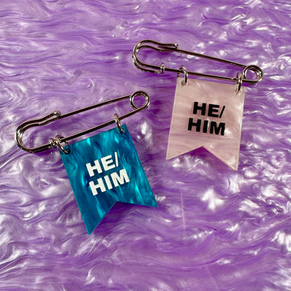Two safety pins with 'HE/HIM' labels on a purple marbled background