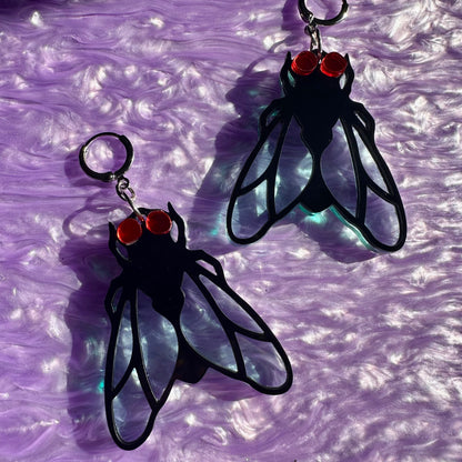 Black fly-shaped earrings with red eyes on a purple textured background