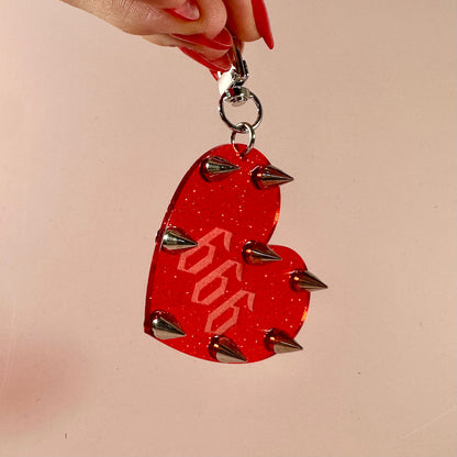 Red heart-shaped keychain with spikes held by a hand against a beige background