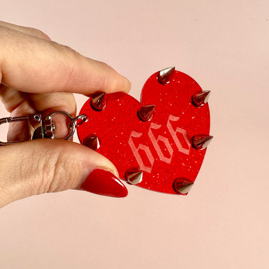 Red heart-shaped keychain with '666' and spikes held by a hand against a pink background