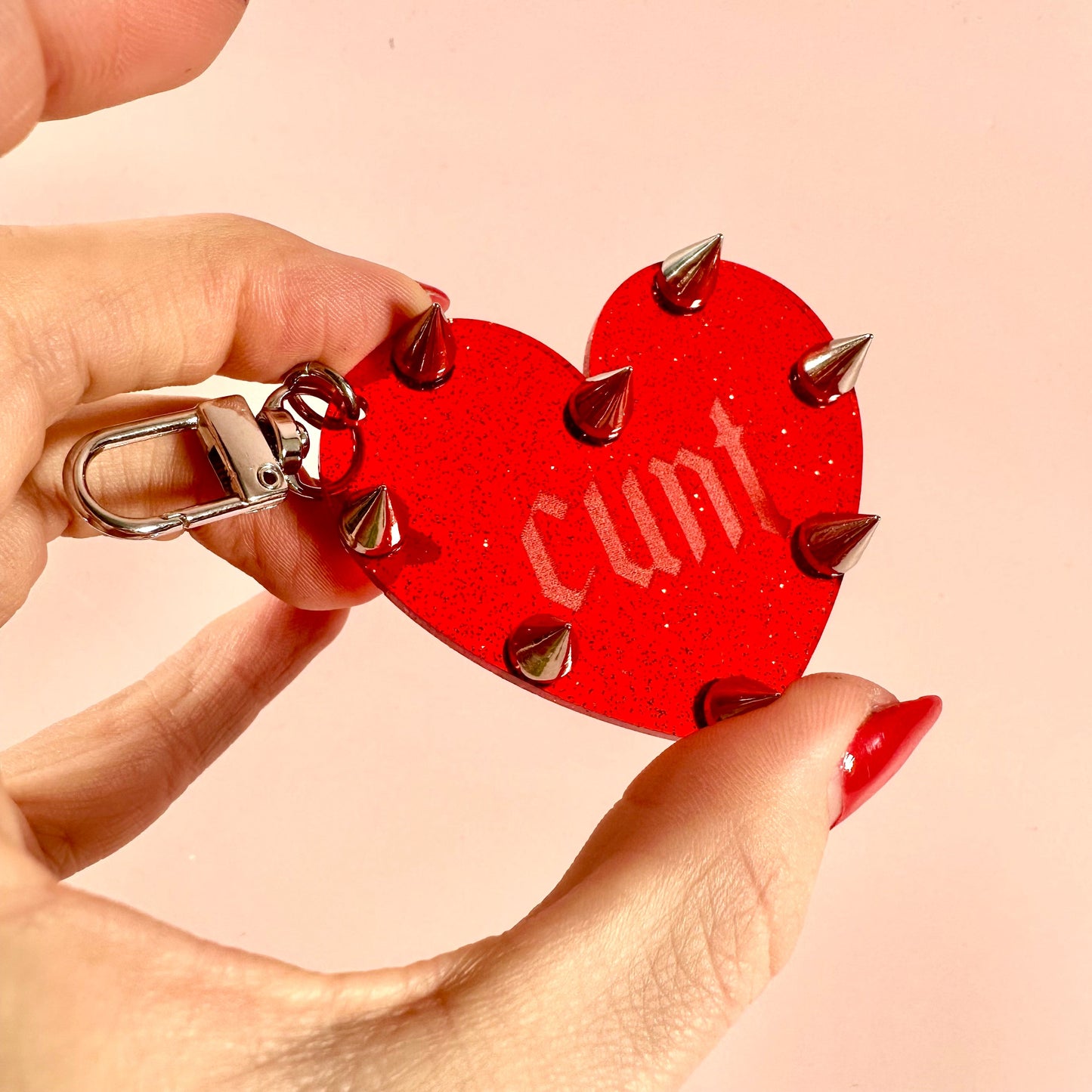 Red heart-shaped keychain with spikes held by a hand against a beige background